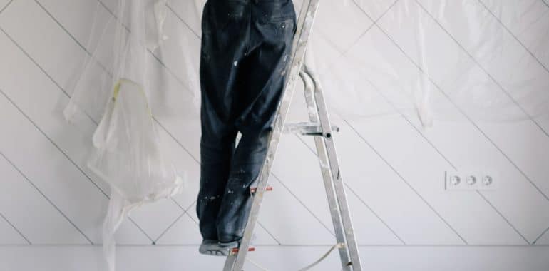 Person in a blue T-shirt standing on a stepladder to install a protective tarpaulin for painting.