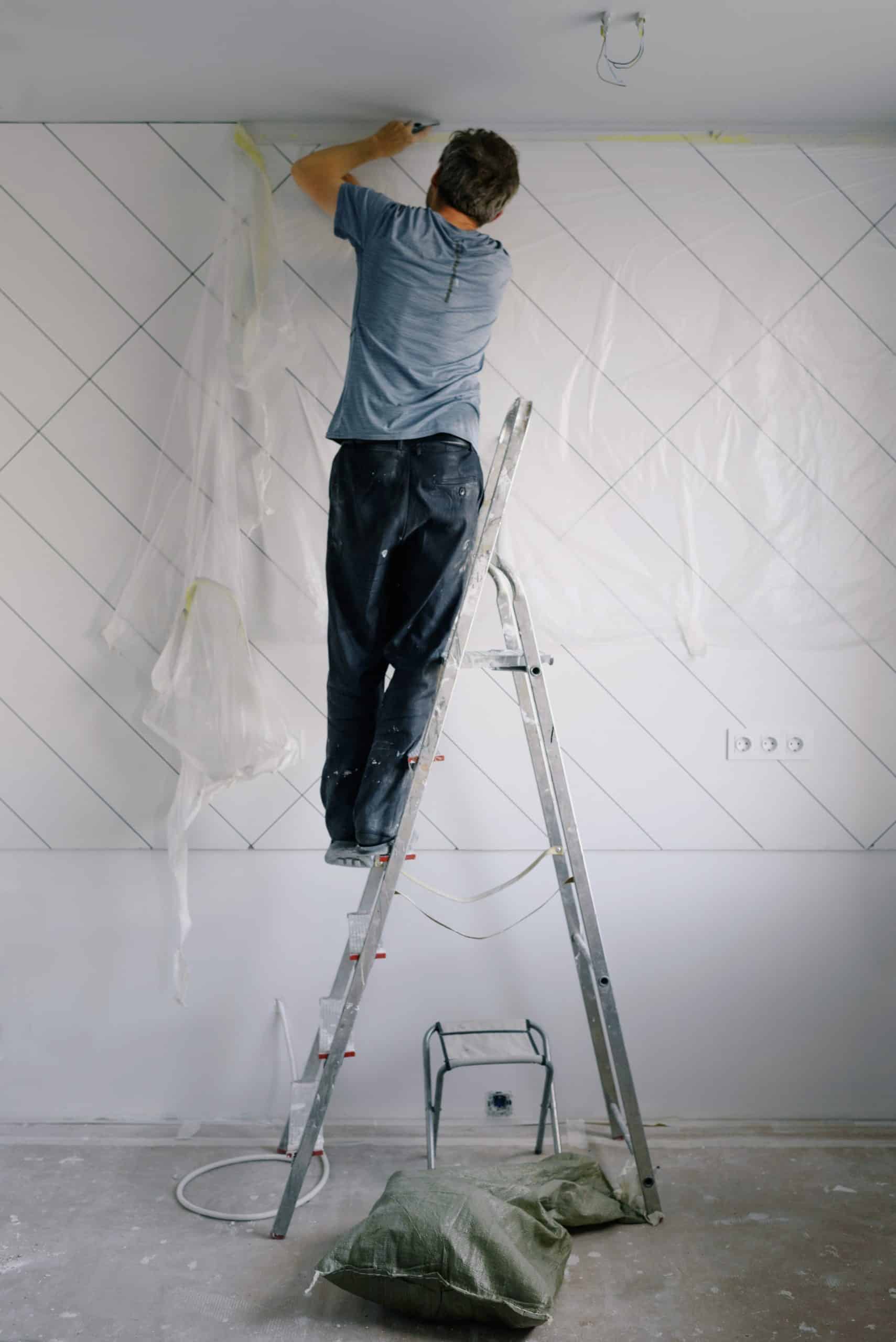 Person in a blue T-shirt standing on a stepladder to install a protective tarpaulin for painting.